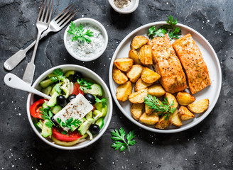 Mediterranean lunch table - baked lemon salmon with potatoes, greek salad, tzadziki sauce on dark background, top view. Flat lay