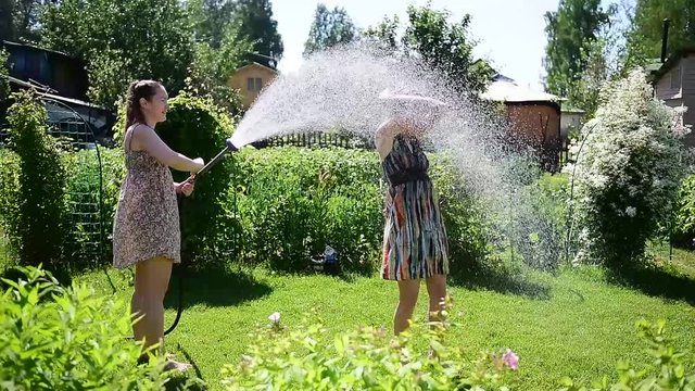 Cute girl pours her girlfriend water from garden hose on  green lawn on a hot summer sunny day. Blonde and brunette in dresses  play and laugh in the yard surrounded by greenery and bright splashes
