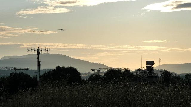 Silhouetted view of an all-around antenna for air defense and tracking made of phased array technology on a rotating platform, with mountains and an aircraft taking off in the background