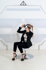 A girl in high heels is sitting in a futuristic room uses virtual reality glasses. Looking forward, holds hand near to the glasses. Full shot. Clear round chair. Spaceship interior. Future