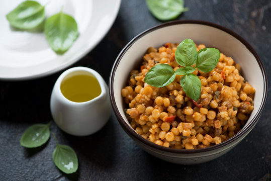 Bowl of fregola pasta with fresh green basil and olive oil on a dark brown stone surface, studio shot