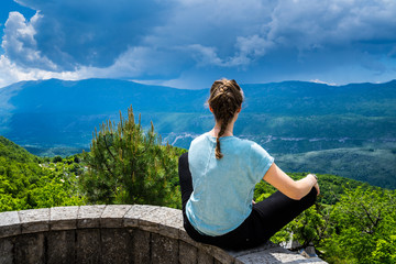 Naklejka na ściany i meble Montenegro, Beautiful young girl sitting in tailor seat on a stone wall enjoying wide view over green nature landscape bjelopavlici plain lowlands valley from mountains near ostrog monastery
