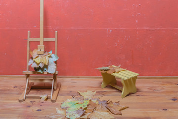 Easel stand with dried leaves, small bench, and dried leaves on floor in studio. Autumn decoration.