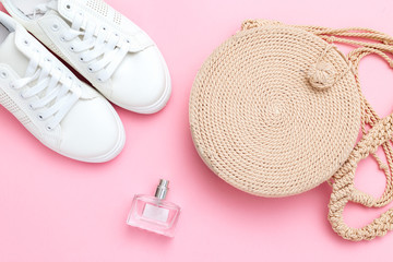 Women's wicker handbag and white sneakers with a perfume on a pink background. Flat lay