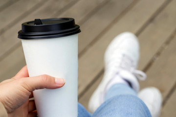 Hand holding white paper cup with black lid, legs with jeans and sneakers background, wooden floor. Take away, coffee shop concept. Close-up, copy space, mock up