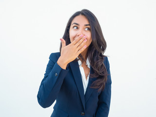 Happy excited office employee learning secret news or gossips. Sly beautiful young Latin woman in office suit covering mouth with hand, looking at copy space away and smiling. Keeping secret concept