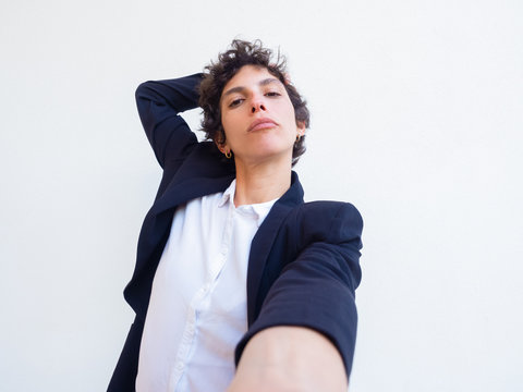 Confident Androgynous Young Woman Holding Cell Phone In Hand And Taking Selfie. Self Portrait Of Female Professional In Office Suit. Poser Concept