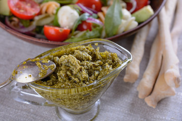 Appetizing pesto sauce in a glass sauce-pan against the background of a bowl with a salad of cherry tomatoes, pasta, arugula and other vegetables next to bread sticks, close up