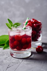 Fresh cherry lemonade with ice, mint and paper straw in sparkling glasses on gray table background, copy space. Cold summer drink. Berry cocktail