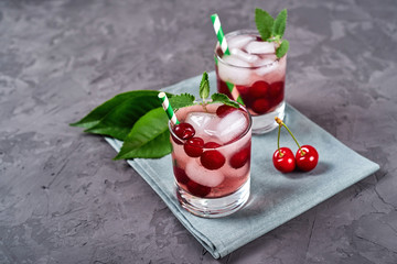 Fresh cherry lemonade with ice, mint and paper straw in sparkling glasses on gray table background, copy space. Cold summer drink. Berry cocktail