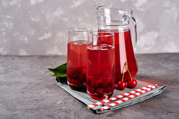Fresh cherry lemonade in sparkling glasses and jug with paper striped straws on gray table background, copy space. Cold summer drink. Berry cocktail