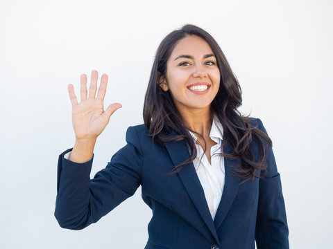Positive friendly businesswoman making greeting gesture. Front view of smiling beautiful young Latin woman in office suit waving hand and saying hello. Gesturing concept