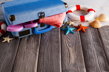 Open holiday suitcase; towel, flip flops, lifebuoy and shells on wooden table and beach