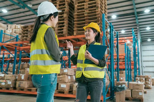Happy female manager and worker standing together in warehouse office. two women staff in storehouse discussing cooperation. young girls employees wearing hard hats and safety vest in stockroom.