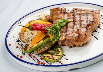 Tender grilled steak or T-bone steak served with grilled vegetables on a white plate.