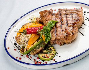 Tender grilled steak or T-bone steak served with grilled vegetables on a white plate.