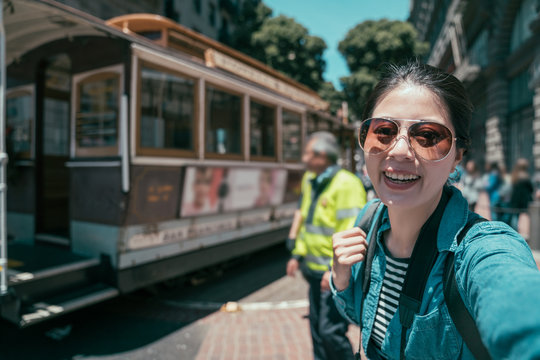 Portrait of young asian woman tourist standing near famous tramway traveling in modern urban in california. smiling lady in sunglasses making self picture against cable car. happy female take selfie