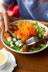 Young woman is having a healthy dietary lunch in a cafe with salad buddha bowl with mixed green salad, grated carrot, cucumbers, nachos, feta and chickpea falafel
