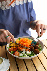 Young woman is having a healthy dietary lunch in a cafe with salad buddha bowl with mixed green salad, grated carrot, cucumbers, nachos, feta and chickpea falafel