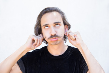 Ridiculous young man with handlebar mustache having fun. Closeup shot of funny brunet holding his ears with hands and making facial expressions. Facial expression concept