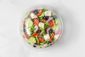 Poster Healthy greek salad in plastic package for take away or food delivery on a white marble background © samael334