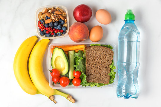 school lunch boxes with sandwich, vegetables, nuts, berries, banana, eggs, peaches and water on white marble background