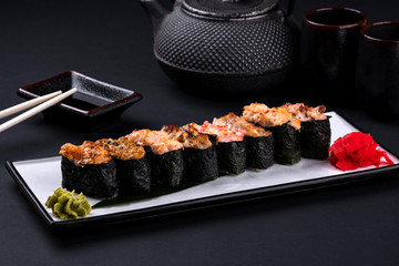 Close up of assorted baked sushi set with salmon, shrimp, caviar, mussels, tuna, cream cheese and hot sauce and chopsticks, soy on black background. Hot spicy rolls and green tea teapot