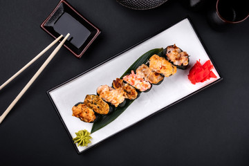 Top view Close up of assorted baked sushi set with salmon, shrimp, caviar, mussels, tuna, cream cheese and hot sauce and chopsticks, soy on black background. Hot spicy rolls and green tea teapot