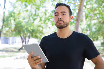 Handsome young man holding tablet computer. Serious bearded man using digital tablet and looking at camera outdoor. Wireless technology concept