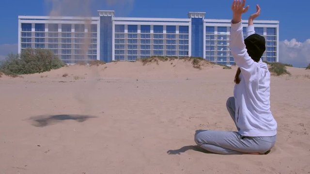 Woman traveller on the beach throws sand that flies in the wind. She rests sitting at sea on sand dunes. Big building on background in coastal city. Girl wears in hat, sweatsirt and trousers.