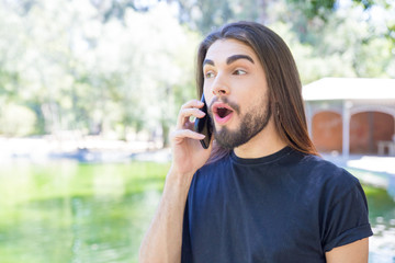 Surprised young man talking on phone in park. Handsome young man with piercing in nose chatting on phone. Communication and technology concept