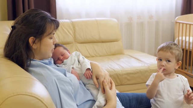 Mother sit on coach holding newborn baby, toddler brother request woman