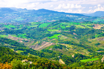 Naklejka na ściany i meble Aerial top panoramic view of landscape with valley, green hills, fields and villages of Republic San Marino suburban district with blue sky white clouds background. View from San Marino fortress
