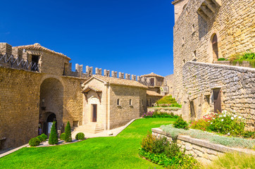 Fototapeta na wymiar Courtyard with green grass lawn of Prima Torre Guaita first medieval tower with stone brick fortress wall with merlons on Mount Titano rock, blue sky white clouds background, Republic San Marino