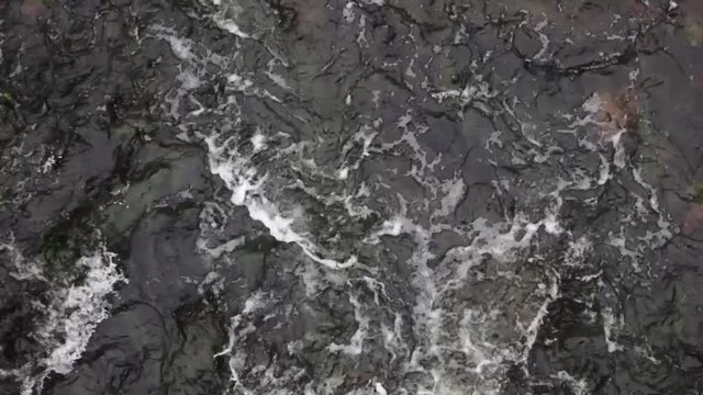 A stream of pure river water rushes between the stones. Waves and splashes of water create a white foam on the surface. Top view of the fast flowing river.