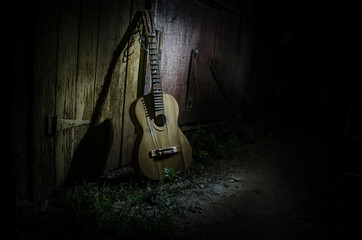 An wooden acoustic guitar is against a grunge textured wall. The room is dark with a spotlight for...