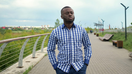 Black guy in plaid shirt and jeans on the street. Young afro american man is walking in city park in cloudy summer day. He folded hands in pockets. He looks worried and anxious.