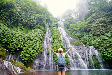 Travel and freedom. Young woman in hat with rucksack enjoying tropical waterfall view.