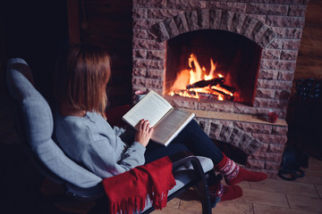 Cozy home. Pretty young woman is reading book near the fireplace.