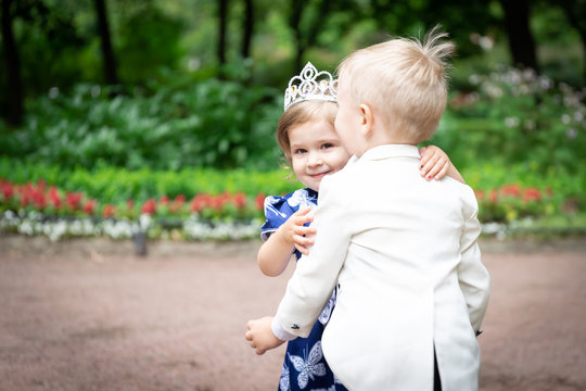 A boy in a dress coat and a girl in a dress against the background of flowers. The boy hugs the girl.