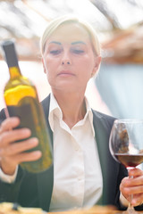 Mature blonde woman in formalwear looking at bottle of cabernet
