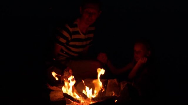 Close-up Of A Man-traveler And A Child By The Fire At The Water In The Summer Outside The City, Dad And Daughter, Dad Straightens Stick Burning Logs In The Fire, The Concept Of Camping And Outdoor Rec