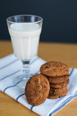 round brown wholemeal oatmeal cookies stack with raisins and white milk in glass cup on striped textile napkin on wooden table, close up side view of vertical still life stock photo image
