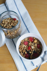granola with fried oatmeal flakes, hazelnuts, peanuts, dried raisins and cherries in glass and white bowl on textile napkin on wooden table, close up top view of vertical still life stock photo image
