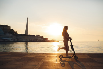 Portrait of a smiling pretty girl in pink clothes, rides on a  scooter and looks at the camera, background city in the sunset. Walk on a kick scooter.Beautiful woman riding a scooter, walking, leisure