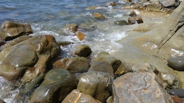 Sea surf on big rocks. Water waves on beach with multicolored polished stones, closeup view. Sunny summer day. With original sound. Natural background.