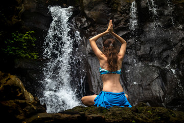 Young Caucasian woman meditating, practicing yoga at waterfall in Ubud, Bali, Indonesia. View from back.