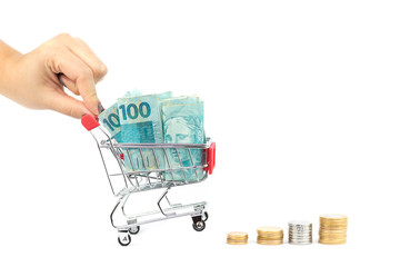 Step of stacks of coins and female hand pushing brazilian notes one hundred (100) reais on shopping cart or supermarket trolley, business finance shopping concept. Brazil money.