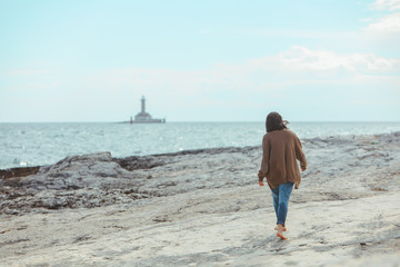 woman walking by rocky sea beach in wet jeans lighthouse on background