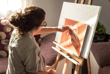 Woman with casual clothes and glasses painting with sanguine or red chalk. Drawing at home in front of a sunny window with easel.